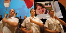 Drei Personen mit Fahnen in den Händen auf einem NPD-Parteitag.