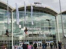 Die Eingangsfassade der Messehallen Hamburg, auf dem Dach haben Menschen ein Banner mit der Aufschrift "Kreuzfahrt stoppen" heruntergelassen. Vor der Halle sind Kletter-Aktivist*innen an Fahnenmasten und eine Mahnwache mit farbenfrohen Anti-Kreuzfahrt-Bannern zu sehen.